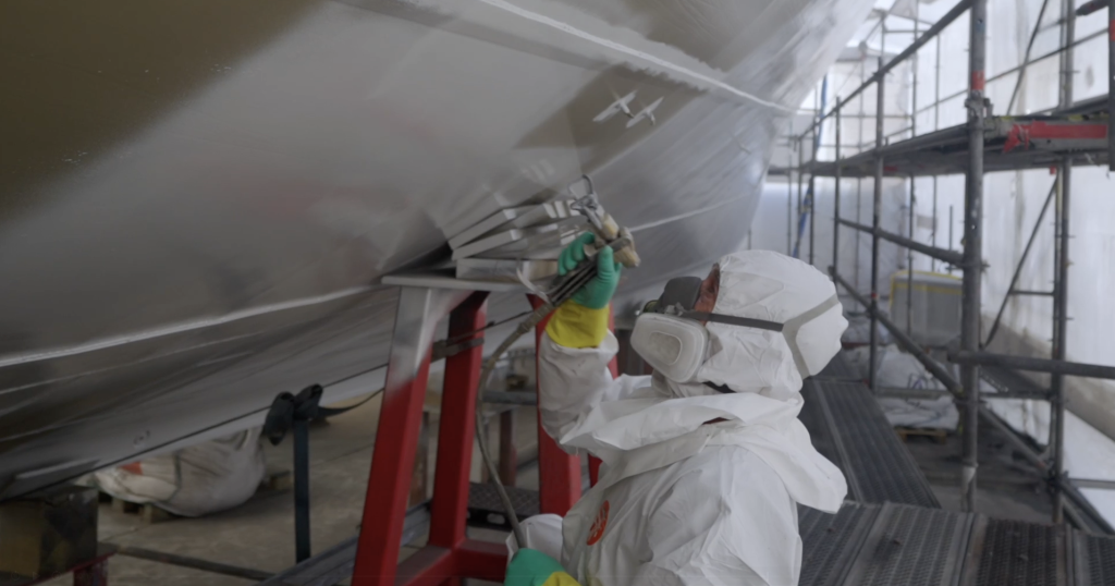Technician applying antifouling paint to the hull of a superyacht in a shipyard using a spray gun with scaffolding in the background